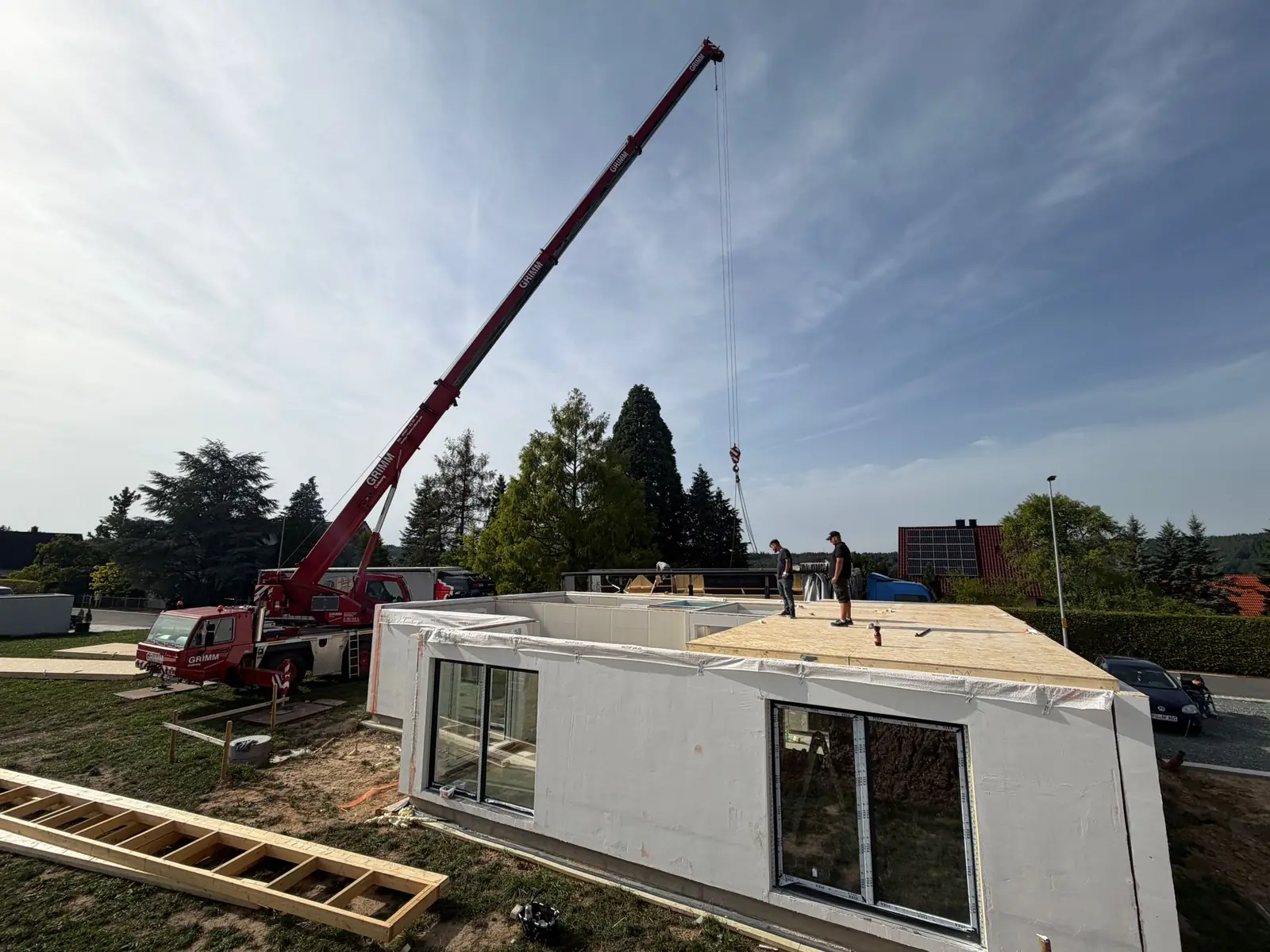 Montage auf der Baustelle: Ein Autokran setzt vorgefertigte Elemente auf die Bodenplatte, zwei Monteure arbeiten auf der Decke